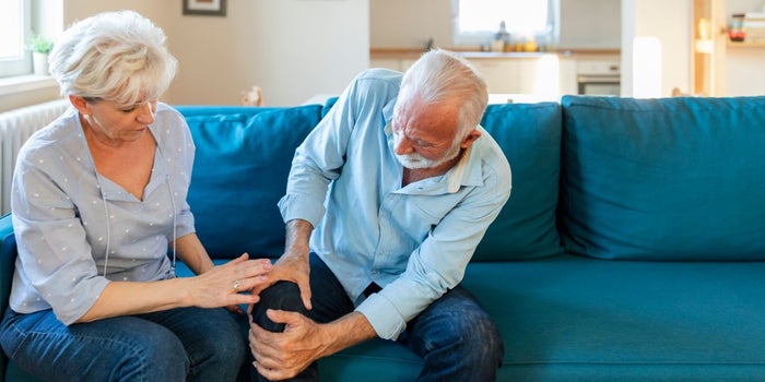 Elderly man suffering from arthritis pain, holding his leg. Elderly woman is checking up on him.